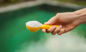 Une main tient une cuillère jaune remplie de poudre blanche, prête à être ajoutée à l'eau verte d'un spa pour un entretien parfait.