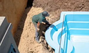 Un ouvrier ajuste une piscine coque bleue dans une excavation. Le terrassement de la piscine coque progresse.