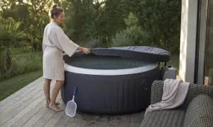 Une femme en peignoir soulève la couverture d'un spa gonflable, prêt pour un moment de détente. Un filet à débris est à proximité, suggérant l'importance de comment entretenir un spa gonflable.