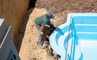 Un ouvrier ajuste une piscine coque bleue dans une excavation. Le terrassement de la piscine coque progresse.