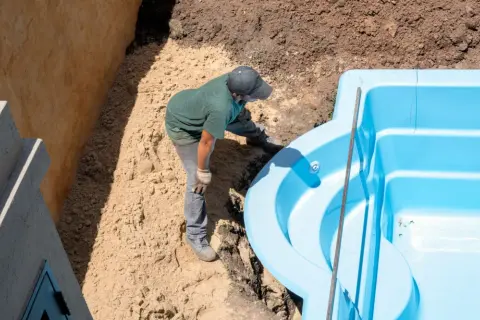 Un ouvrier ajuste une piscine coque bleue dans une excavation. Le terrassement de la piscine coque progresse.