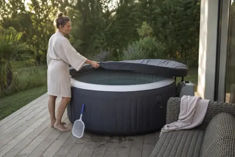 Une femme en peignoir soulève la couverture d'un spa gonflable, prêt pour un moment de détente. Un filet à débris est à proximité, suggérant l'importance de comment entretenir un spa gonflable.