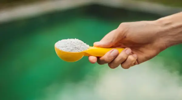 Une main tient une cuillère jaune remplie de poudre blanche, prête à être ajoutée à l'eau verte d'un spa pour un entretien parfait.
