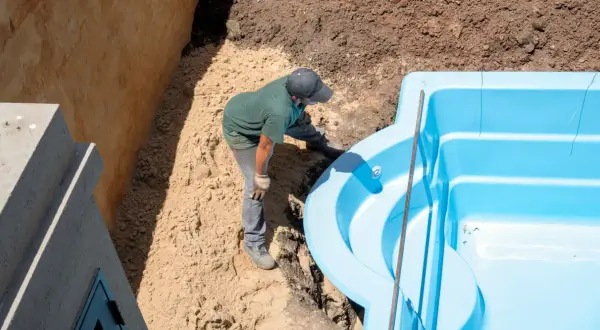 Un ouvrier ajuste une piscine coque bleue dans une excavation. Le terrassement de la piscine coque progresse.