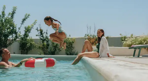 Une famille profite d'une journée ensoleillée à la piscine. Une jeune fille saute dans l'eau, son père la regarde avec un sourire, et sa sœur est assise au bord. Le taux de sel piscine est parfait pour cette baignade joyeuse.