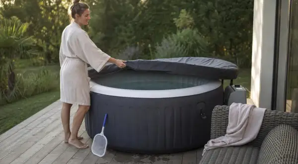 Une femme en peignoir soulève la couverture d'un spa gonflable, prêt pour un moment de détente. Un filet à débris est à proximité, suggérant l'importance de comment entretenir un spa gonflable.