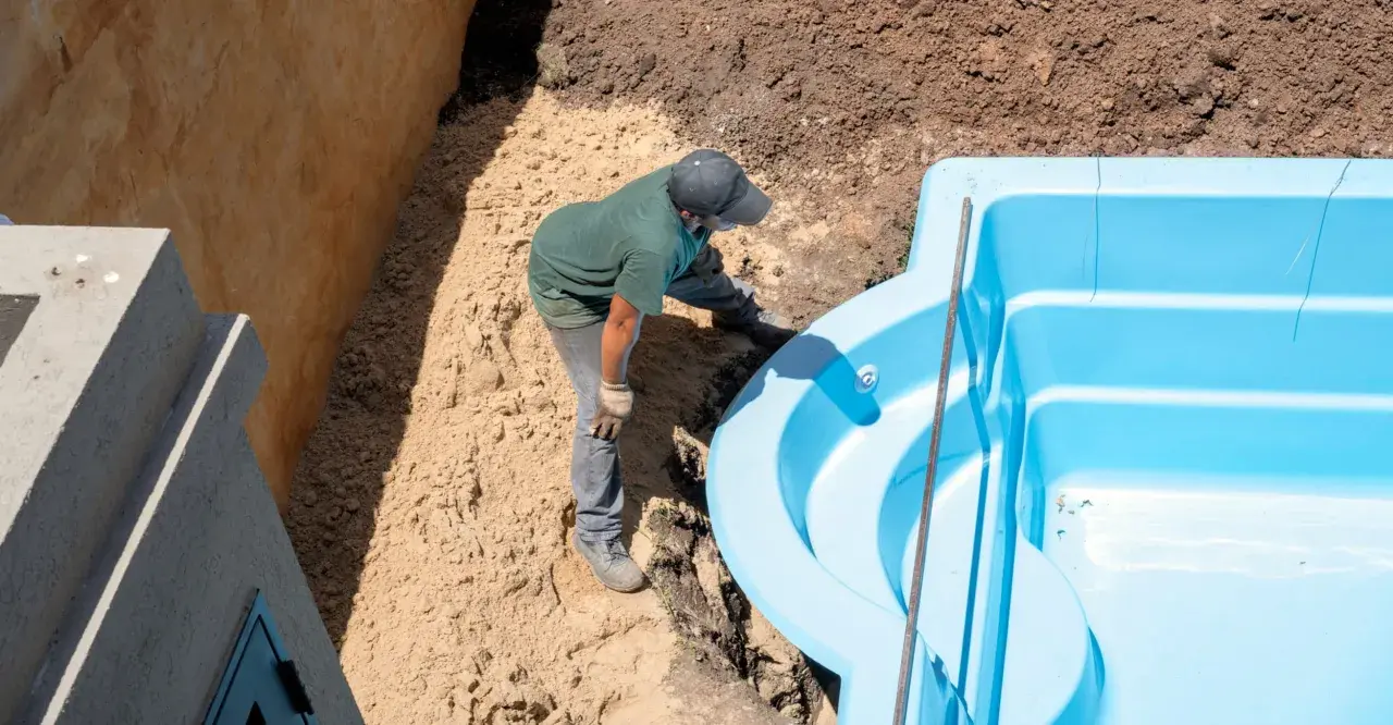 Un ouvrier ajuste une piscine coque bleue dans une excavation. Le terrassement de la piscine coque progresse.