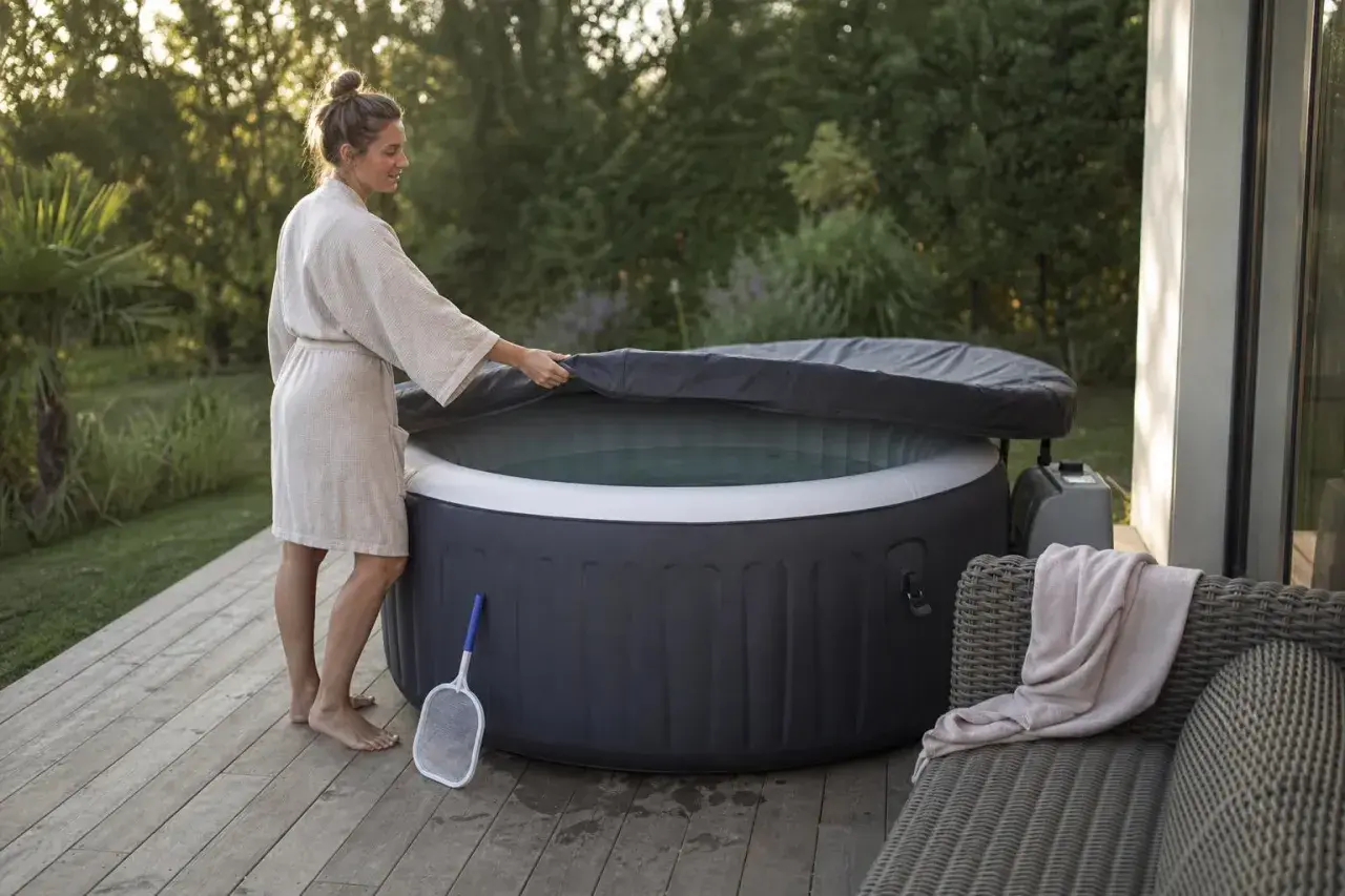 Une femme en peignoir soulève la couverture d'un spa gonflable, prêt pour un moment de détente. Un filet à débris est à proximité, suggérant l'importance de comment entretenir un spa gonflable.