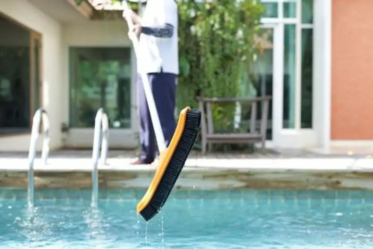 Une brosse de piscine, tenue par une personne, sort de l'eau. C'est un guide d'entretien piscine pour les nuls.