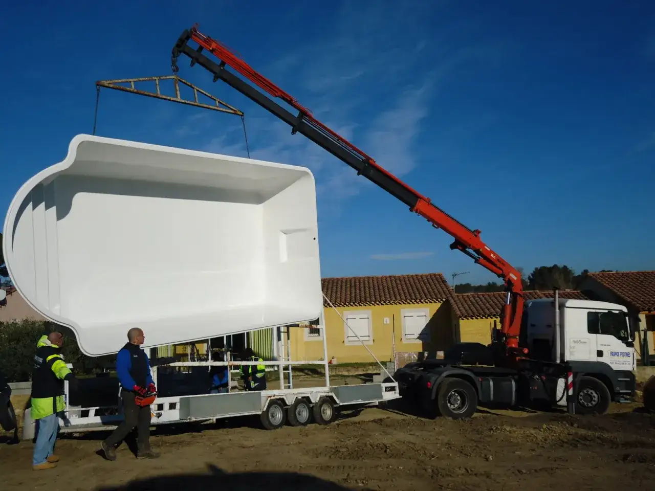 Grue soulevant une piscine coque blanche pour son terrassement. Des ouvriers supervisent l'opération.