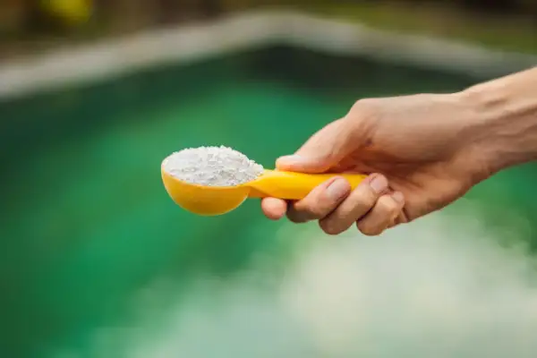 Une main tient une cuillère jaune remplie de poudre blanche, prête à être ajoutée à l'eau verte d'un spa pour un entretien parfait.